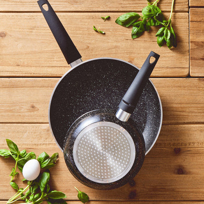 Black frying pan with a perforated bottom on a wooden surface with basil leaves.