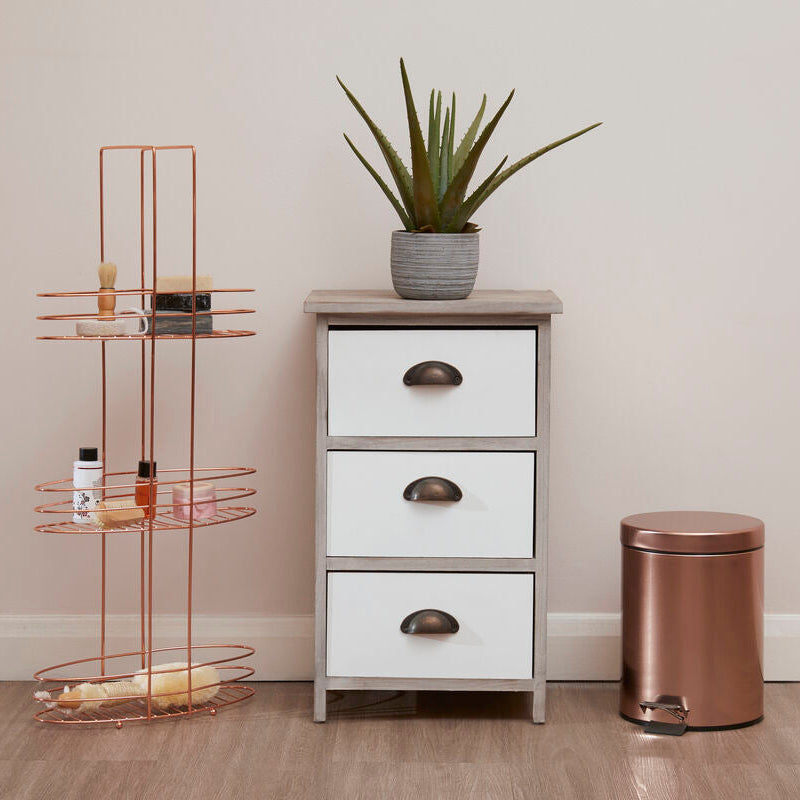White dresser with three drawers, a bronze trash can, and a plant against a plain wall.