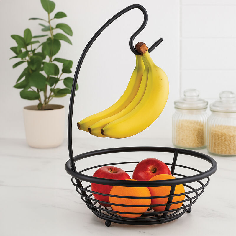 Black wire fruit basket with bananas and apples on a white surface with a plant and jars in the background.