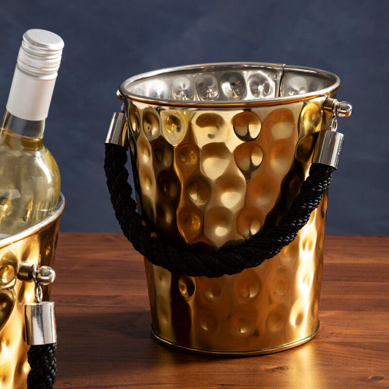 Gold ice bucket with black rope handle on a wooden surface next to a bottle of white wine.