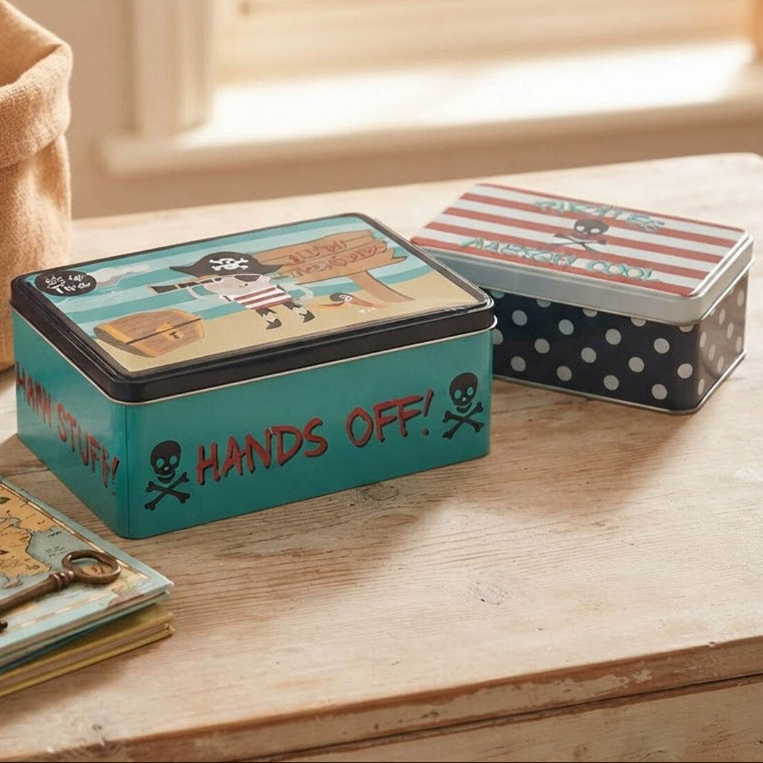 Two pirate-themed metal storage boxes displayed on a wooden table. The box in the foreground is teal with red text reading &