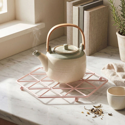 A cream-colored ceramic teapot with a light green lid and a rose gold metal handle, resting on a pink geometric silicone trivet on a white marble countertop. A small matching cream cup sits nearby, with books and a potted plant in the background.