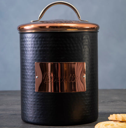 Black cookie jar with copper lid and accents on a wooden surface with cookies.