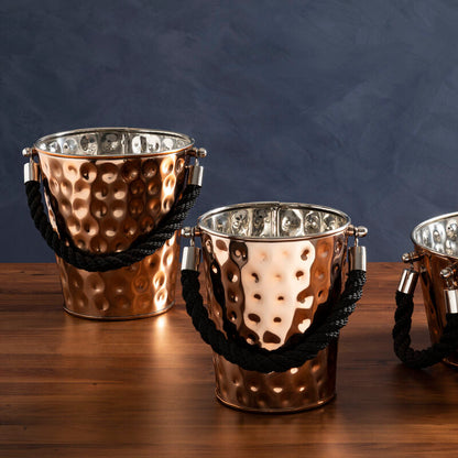 Three copper-colored metal buckets with handles on a wooden surface against a dark background