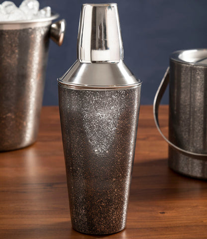Silver cocktail shaker on a wooden surface with ice buckets in the background.