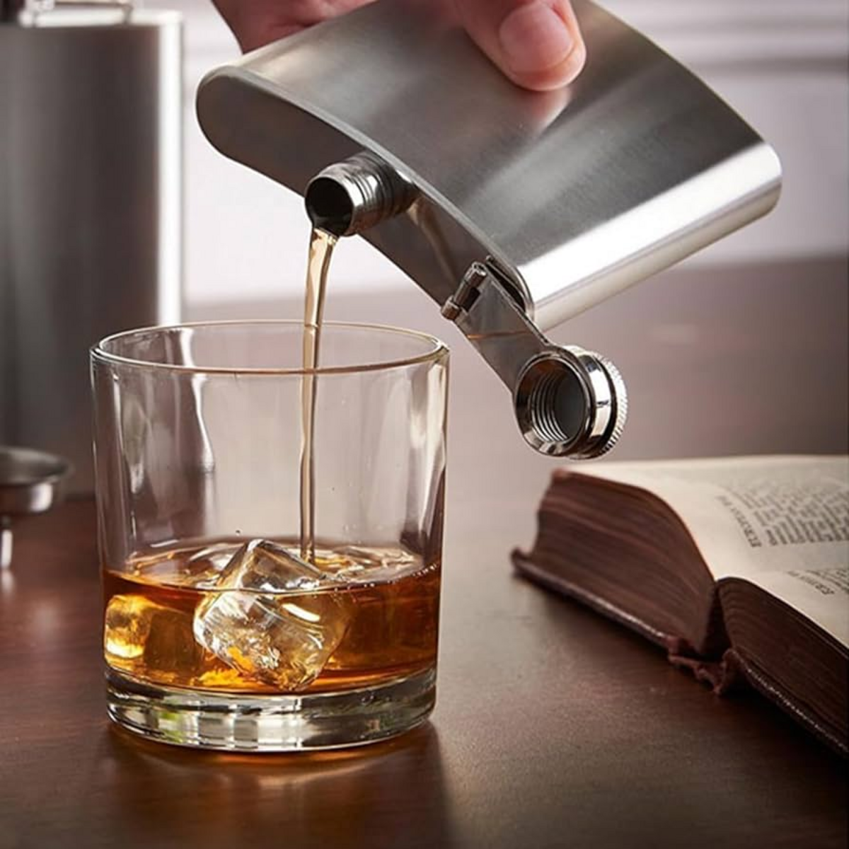 Person pouring whiskey from a metal flask into a glass with ice on a wooden table.