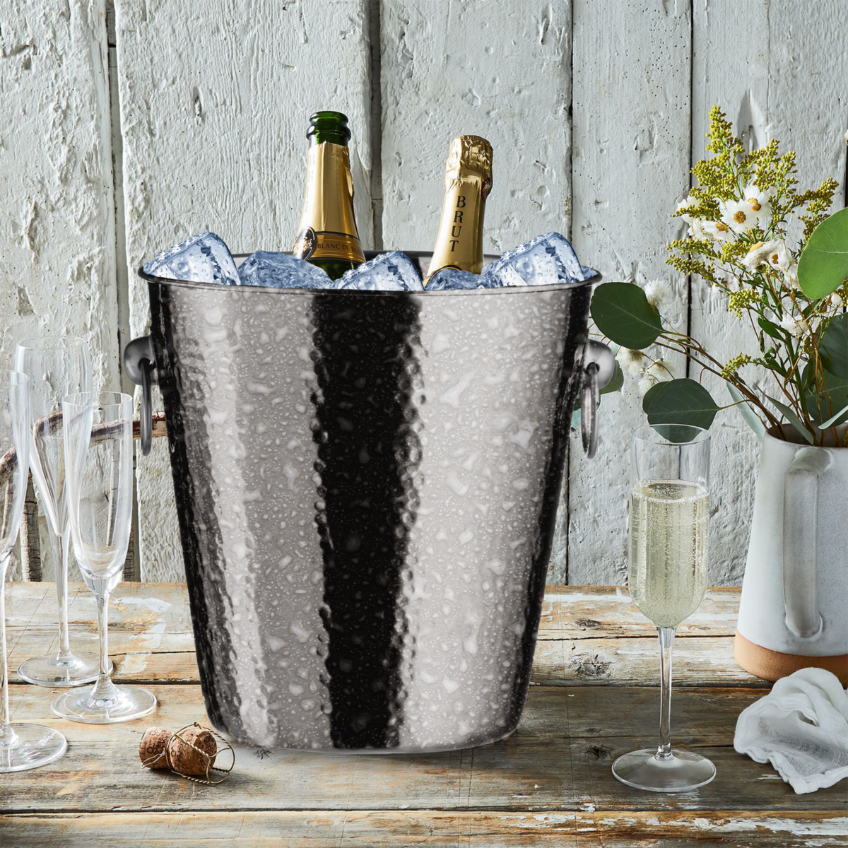 Hammered metal ice bucket with champagne bottles on a rustic wooden table.