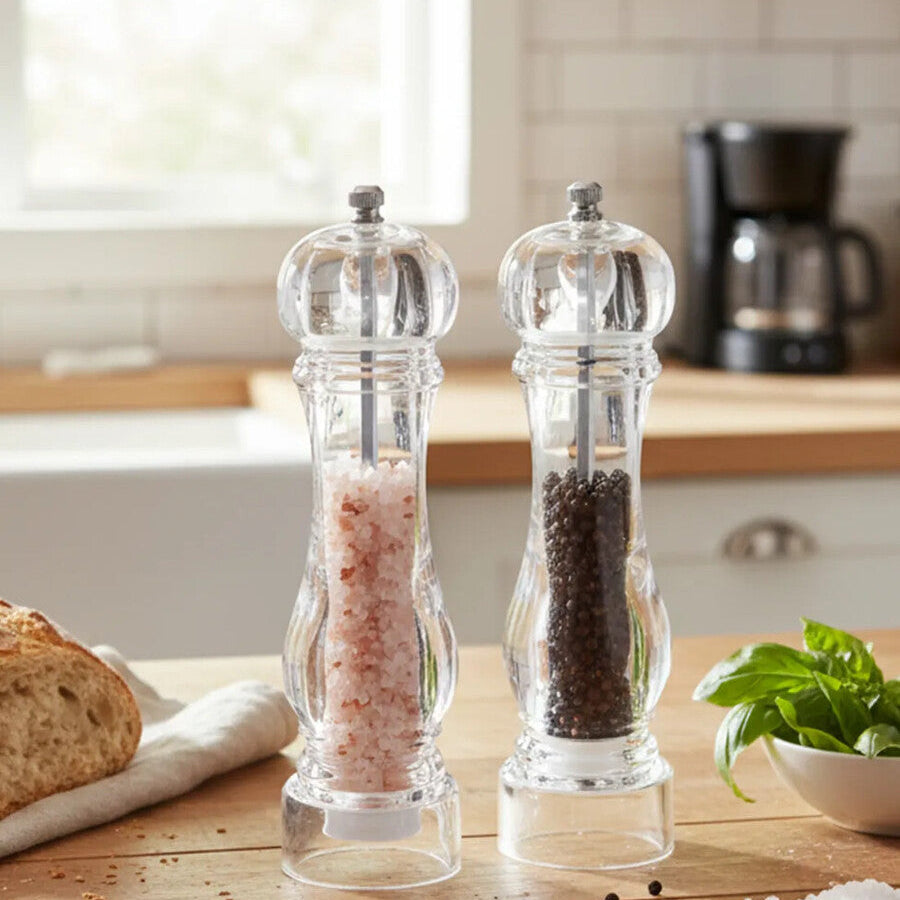 Two pepper grinders on a kitchen counter with bread and herbs in the background.