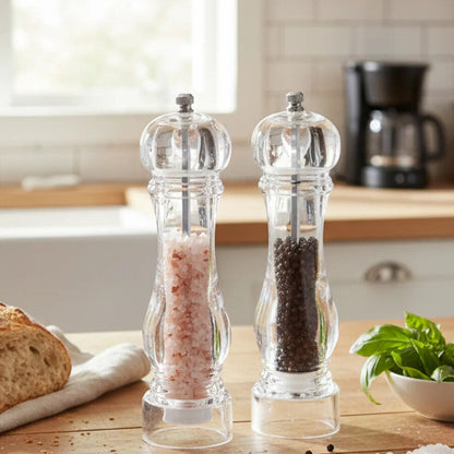 Two pepper grinders on a kitchen counter with bread and herbs in the background.
