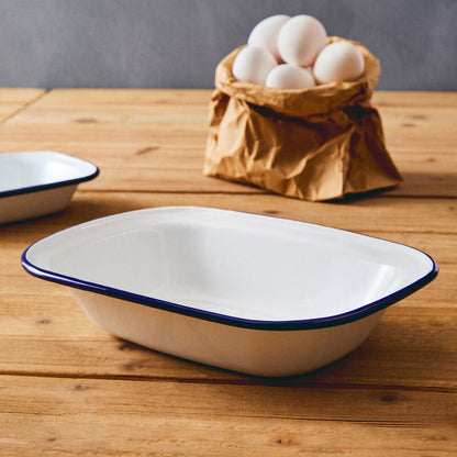 White enameled baking dish with blue rim on a wooden surface, with eggs in a brown paper bag in the background.