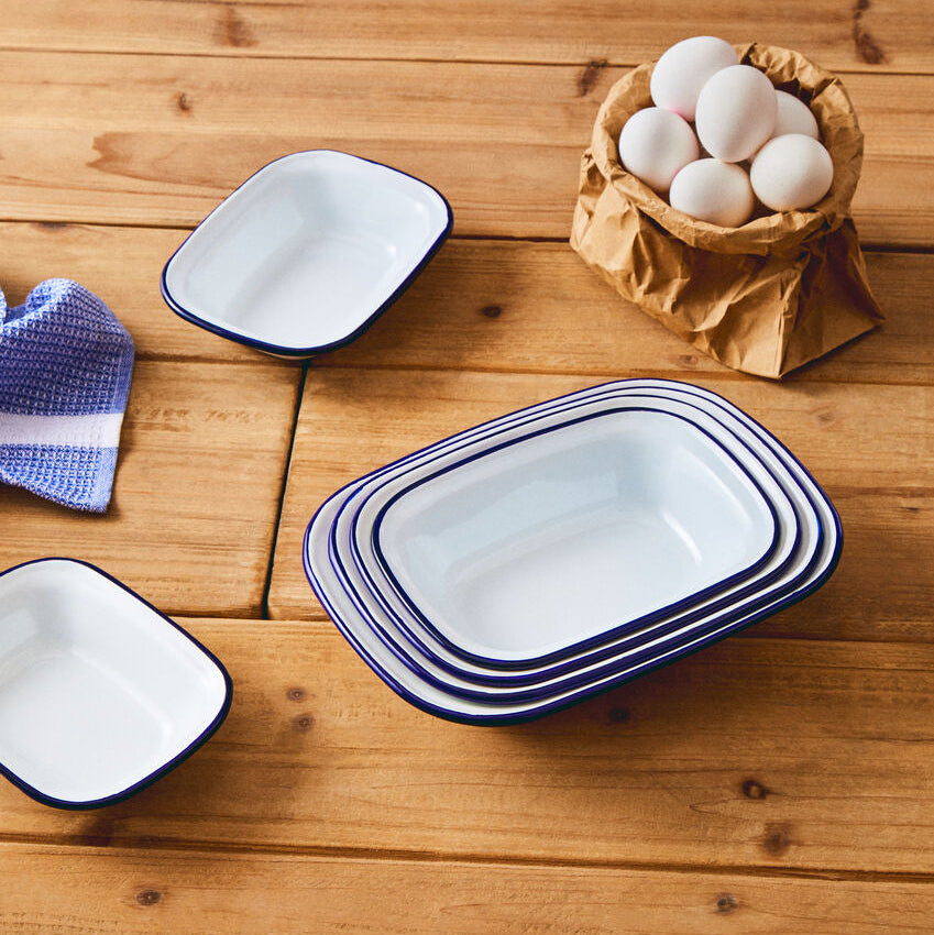 Set of white enameled cookware with blue rims on a wooden surface with eggs in a paper bag.