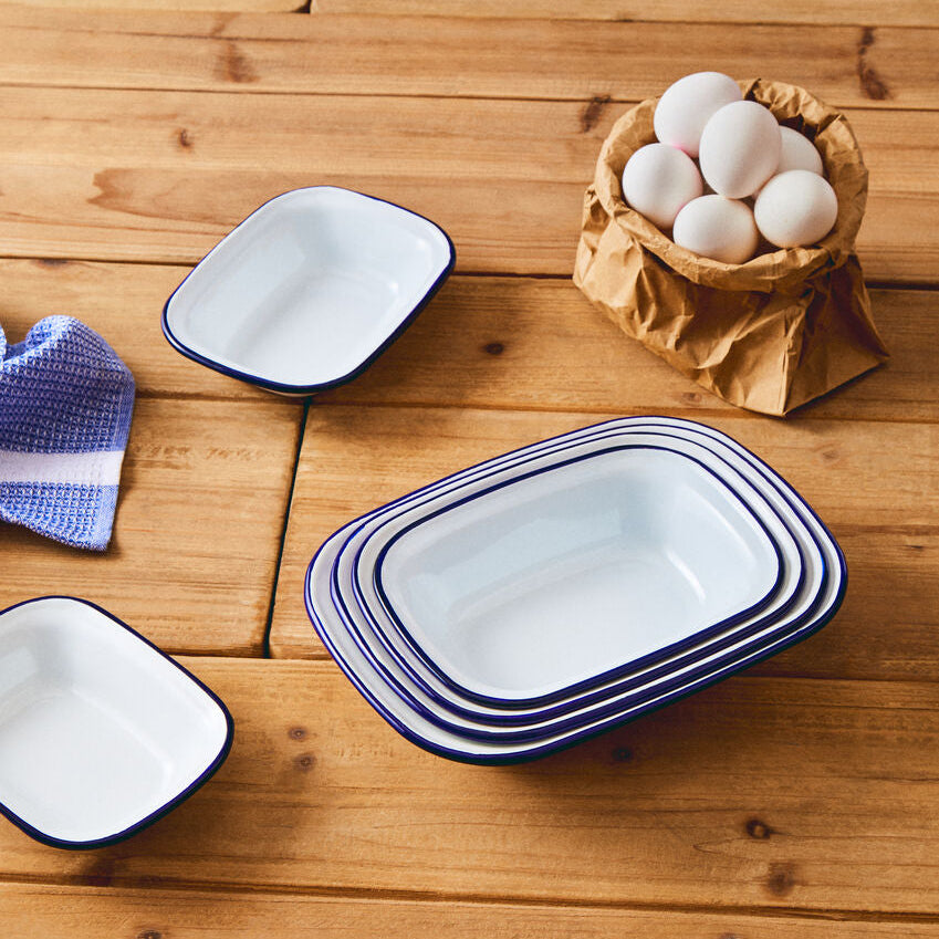 Set of white enameled cookware with blue rims on a wooden surface with eggs in a paper bag.