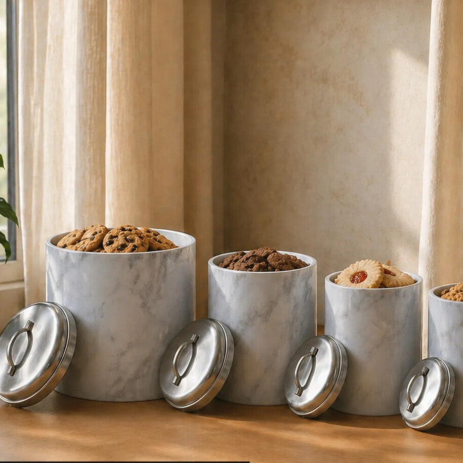 Four white marble finish canisters with silver lids on a wooden surface near a window.