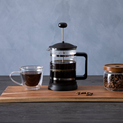French press, glass of coffee, jar of coffee beans on a wooden board with a gray background