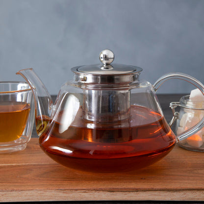 Glass teapot with a silver lid on a wooden surface, filled with tea.