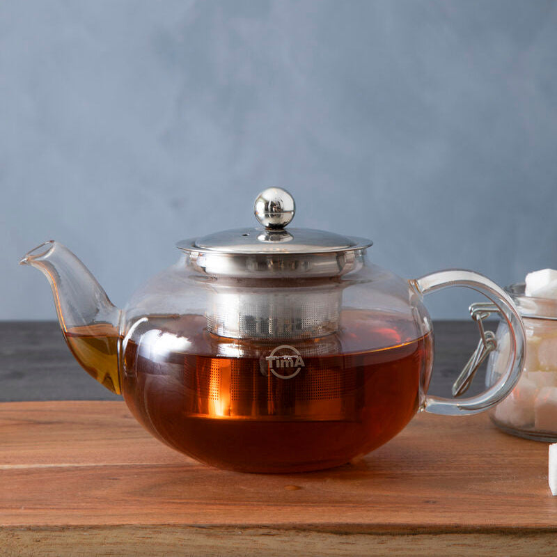 Clear glass teapot with a silver lid on a wooden surface against a gray background