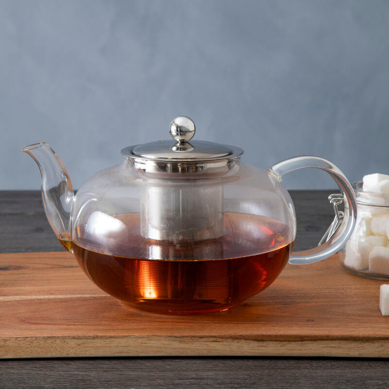 Clear glass teapot with metal lid on a wooden board against a gray background