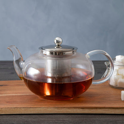 Clear glass teapot with metal lid on a wooden board against a gray background