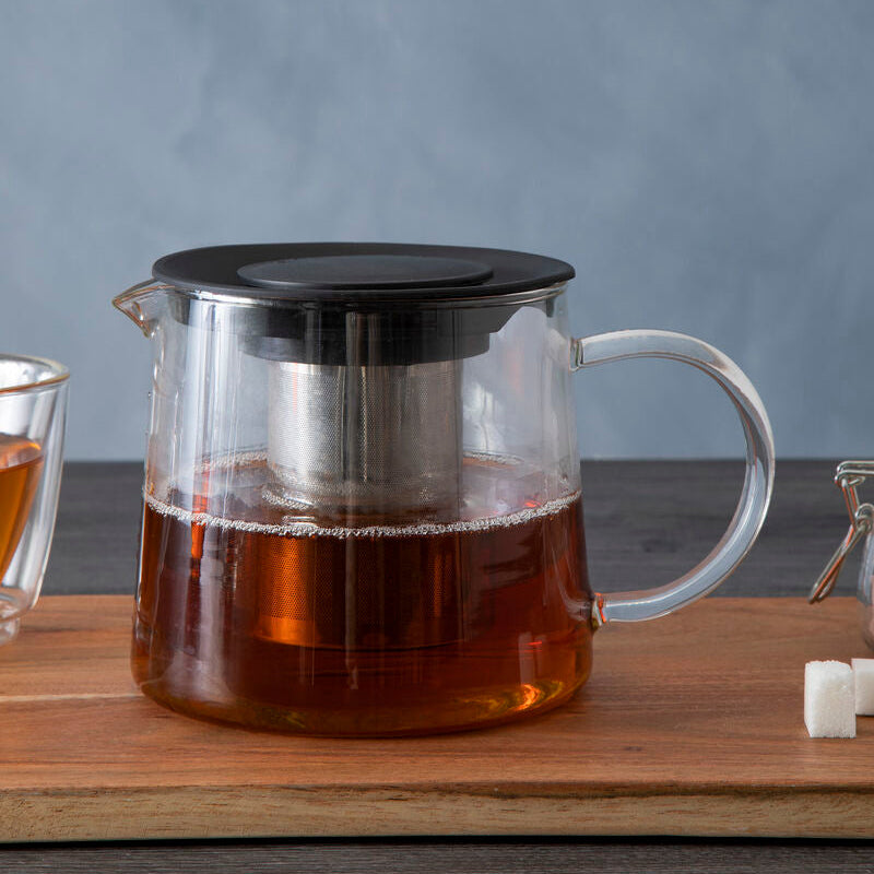 Clear glass teapot with black lid on a wooden surface, with a gray background