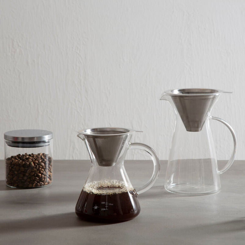 Coffee-making setup with coffee beans, coffee filter, glass carafe, and clear cup on a gray surface.