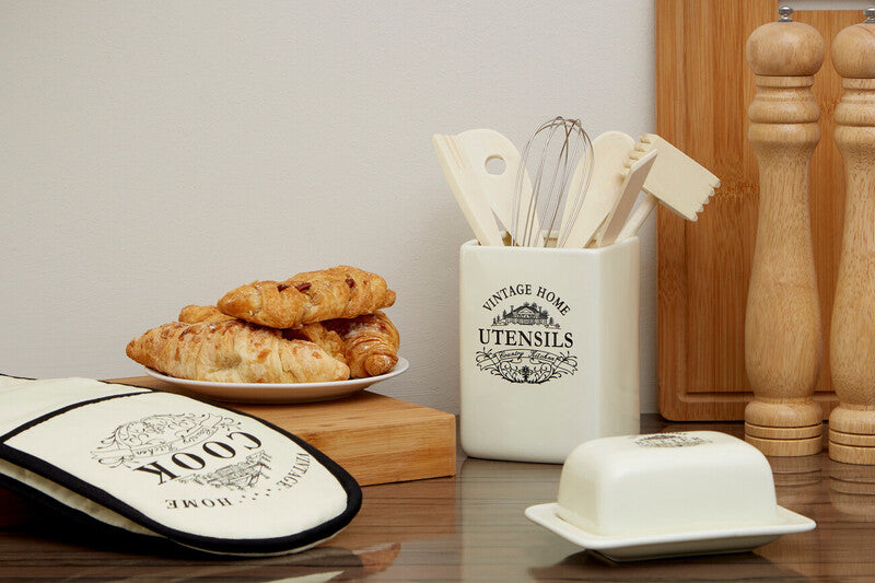 Kitchen utensils set in a container with bread and a cutting board on a wooden surface.