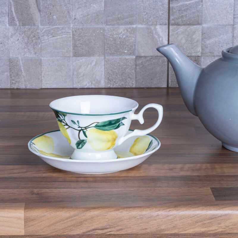 Teacup with lemon design on a saucer next to a gray teapot on a wooden surface with a tiled wall background.