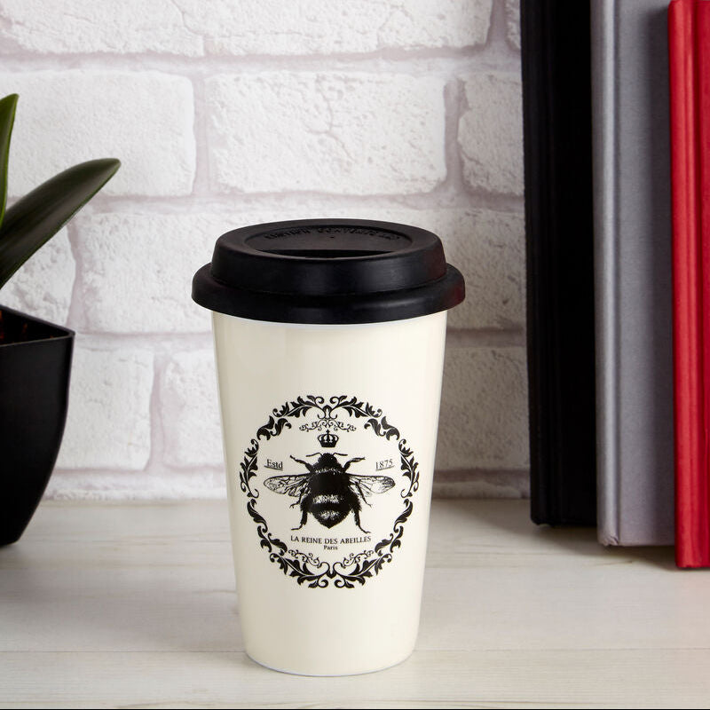 White coffee cup with black lid and bee design on a white surface with books and flowers.