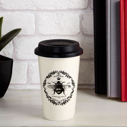 White coffee cup with black lid and bee design on a white surface with books and flowers.