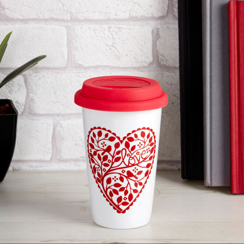 White mug with red floral heart design and red lid on a white surface with books and flowers in the background.