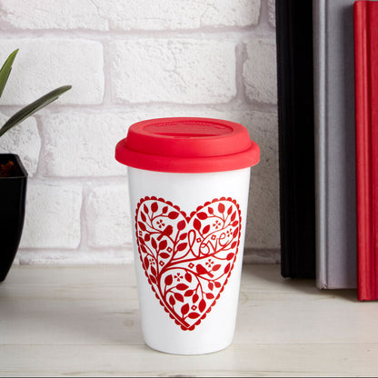 White mug with red floral heart design and red lid on a white surface with books and flowers in the background.