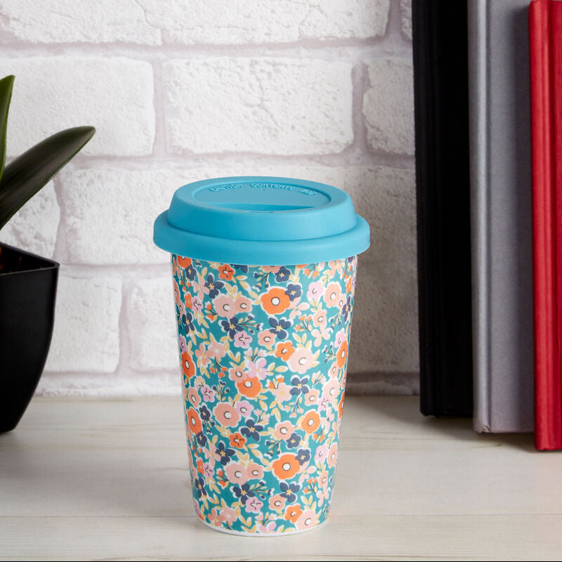 Reusable coffee cup with floral design and blue lid on a white surface with books and flowers in the background.