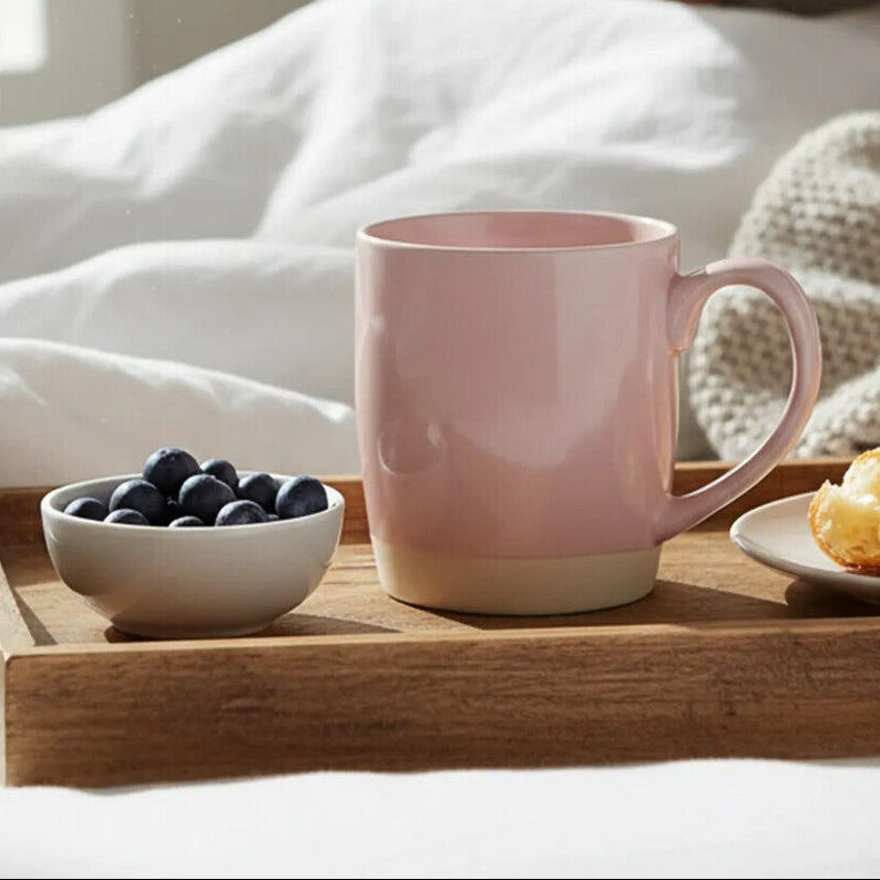 Person reading a book with a pink mug, bowl of blueberries, and plate of pastries on a wooden tray.