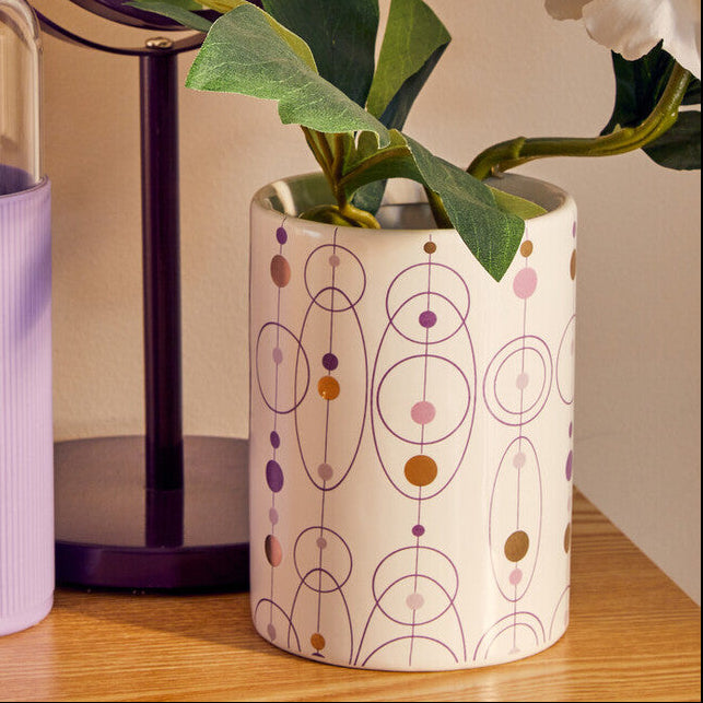 Clear water bottle with purple sleeve, small plant in a decorative pot, and round mirror on a wooden surface.