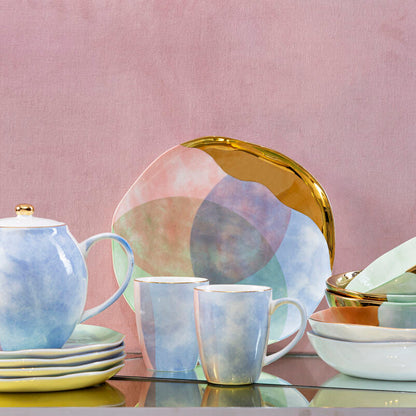 Ceramic tableware set with teapot, mugs, and plates on a reflective surface with a pink background