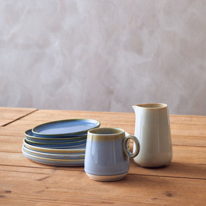 Ceramic tableware including a mug, plates, and a pitcher on a wooden surface with a neutral background.