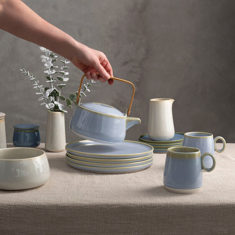 Ceramic tableware set on a neutral background with a hand reaching for a teapot.