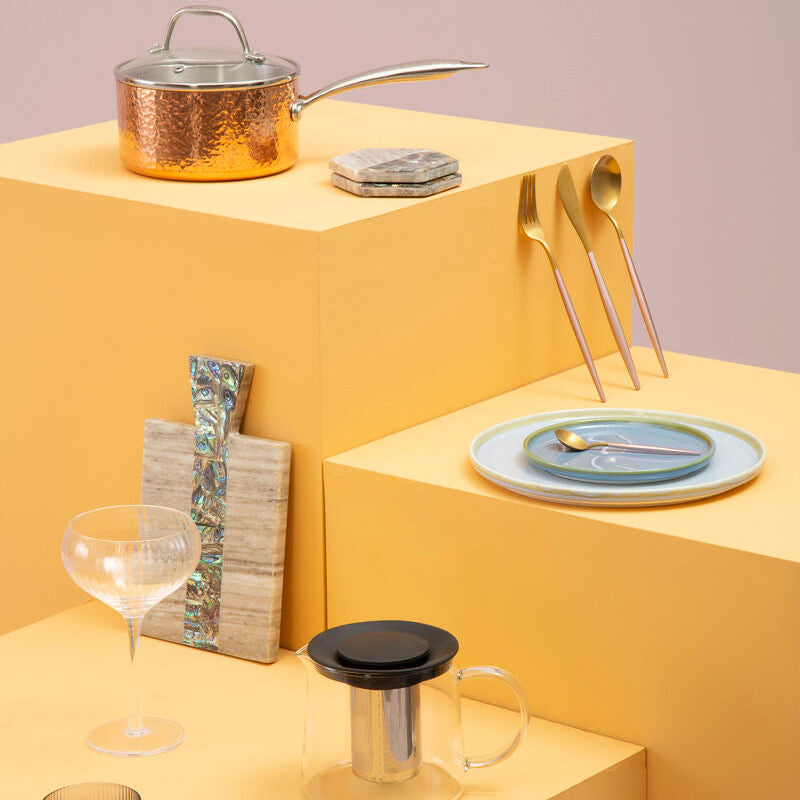 Table setting with a pot, plates, cutlery, and glasses on a yellow platform against a light purple background.