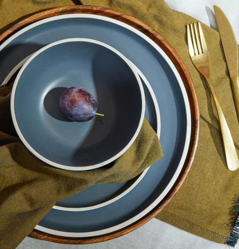 Blue ceramic plates with plums on a green cloth, gold fork, and knife.