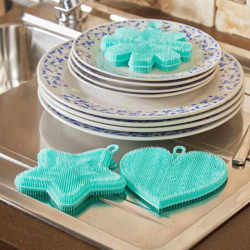 Stack of plates with decorative edge, utensils, and green dish scrubbers on a kitchen counter.