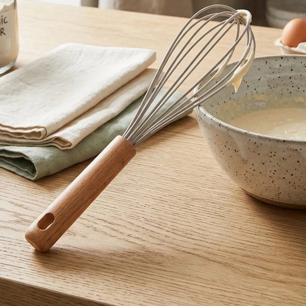 Wooden-handled whisk on a wooden surface with a bowl of batter and flour container in the background.