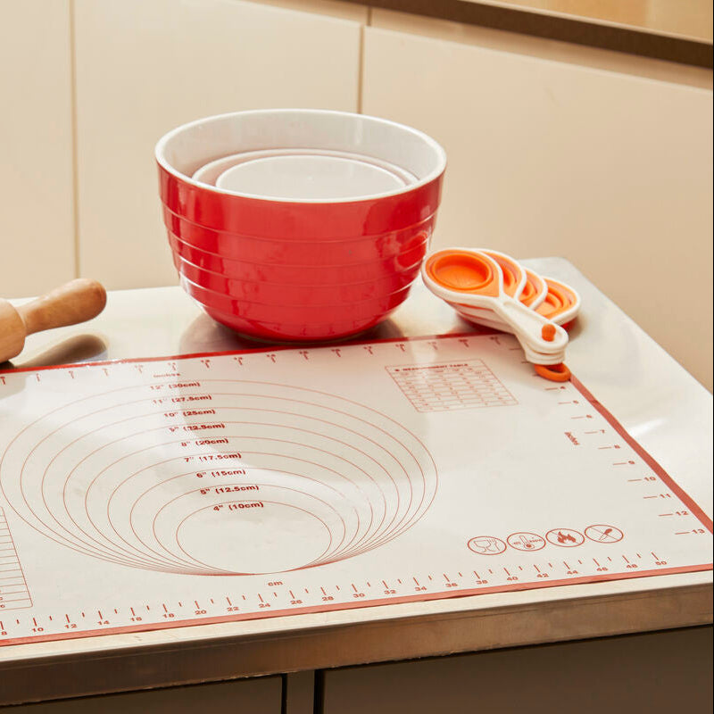 Red mixing bowl, measuring cups, and rolling pin on a baking mat in a kitchen setting.