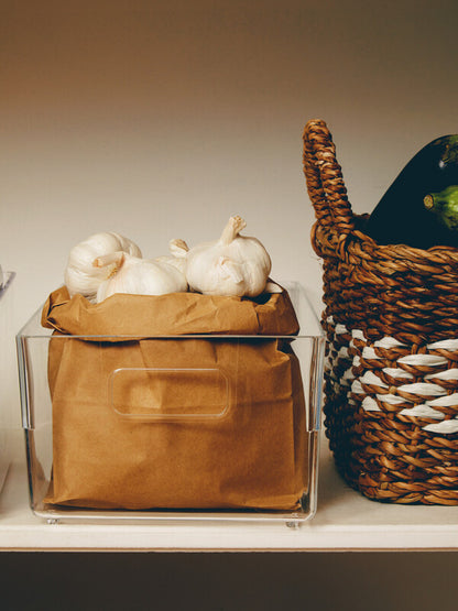 Clear container with brown fabric and garlic bulbs next to a wicker basket on a neutral background