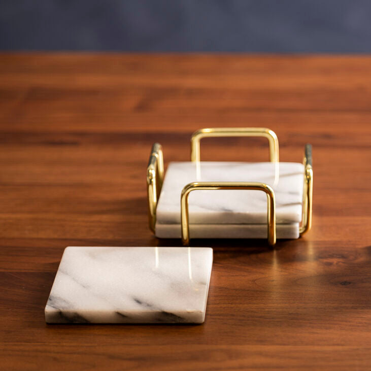 Glass with a drink on a marble coaster, next to a gold holder and marble coasters on a wooden table.