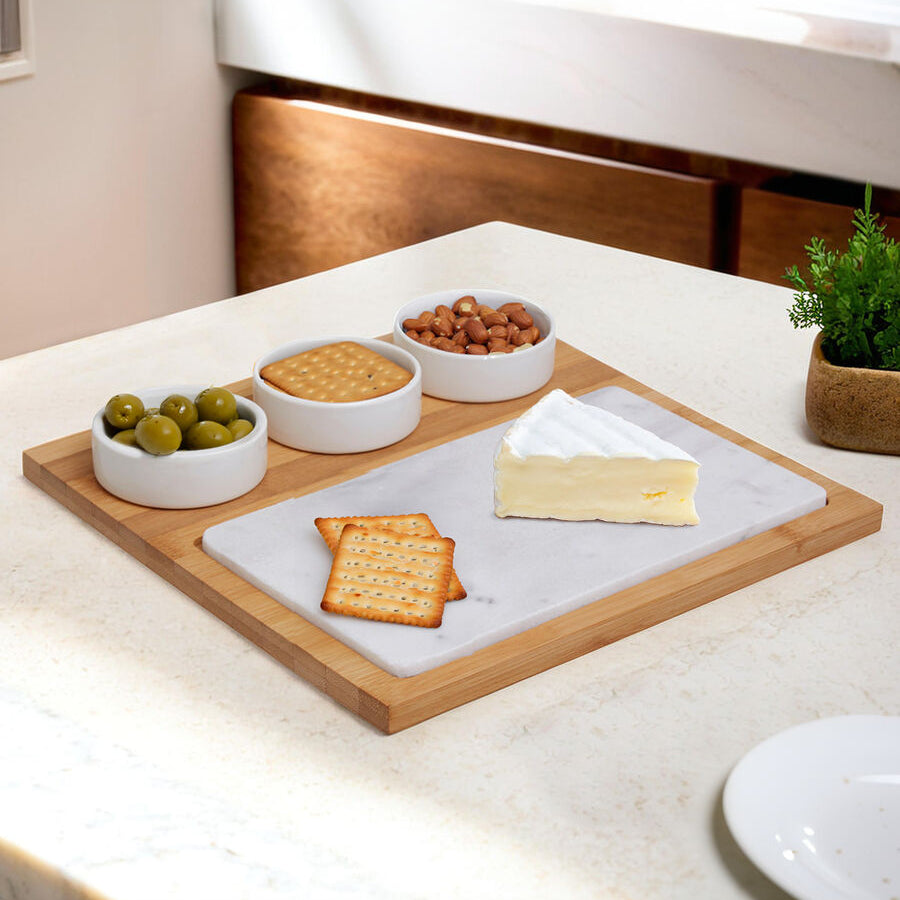 Wooden tray with marble base holding cheese, crackers, and small bowls of olives and nuts on a kitchen counter.