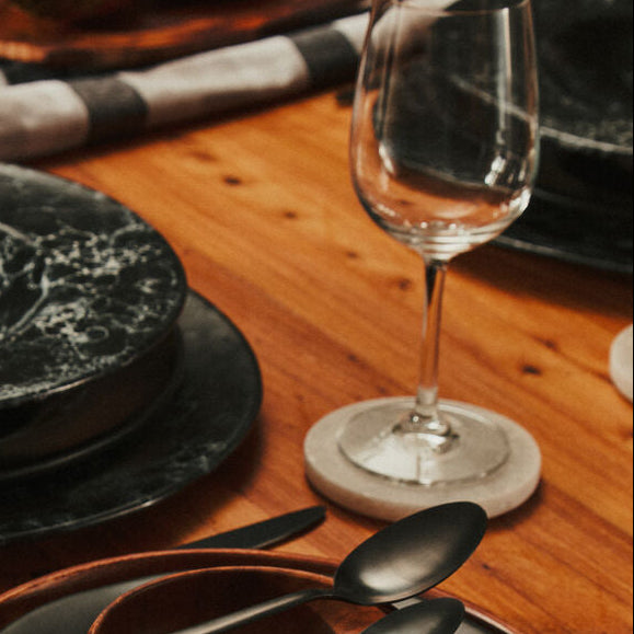 Dining table setting with black plates, wooden bowl, and silverware on a wooden surface.