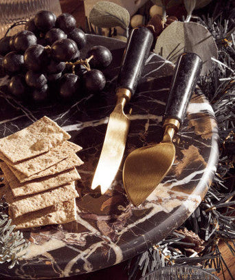 Marble cheese board with crackers, grapes, and gold cheese knives on a festive background.