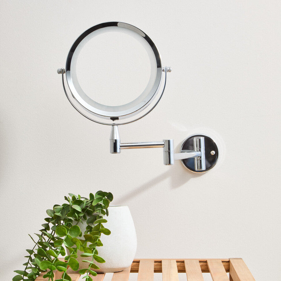 Wall-mounted makeup mirror with a plant and vase on a wooden stool against a white wall.