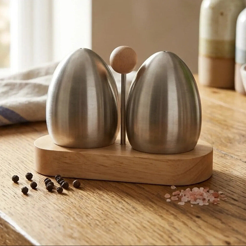 Polished metal salt and pepper shakers on a wooden base with visible ingredients on a kitchen counter.