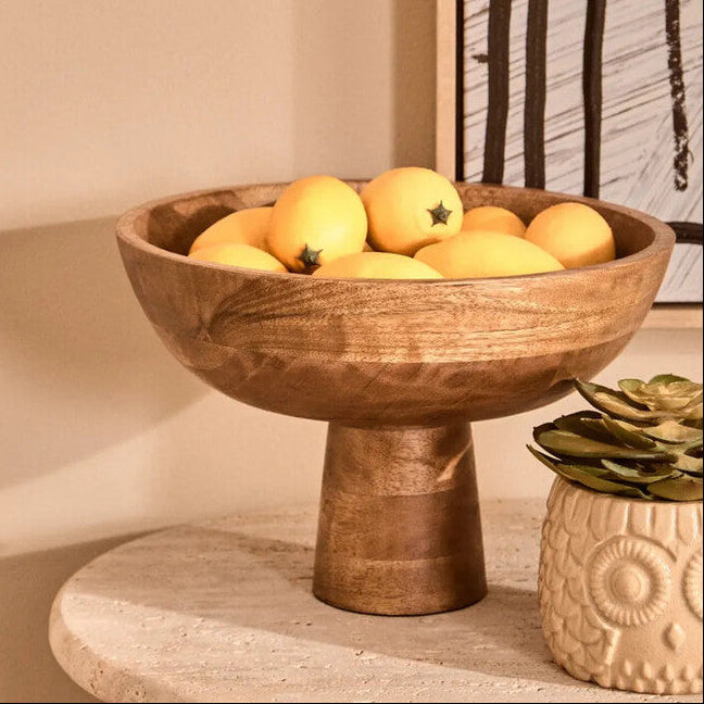 Wooden bowl with lemons on a table next to a small plant, with abstract art in the background.
