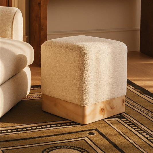 Beige cube-shaped ottoman with wooden base in a room setting.